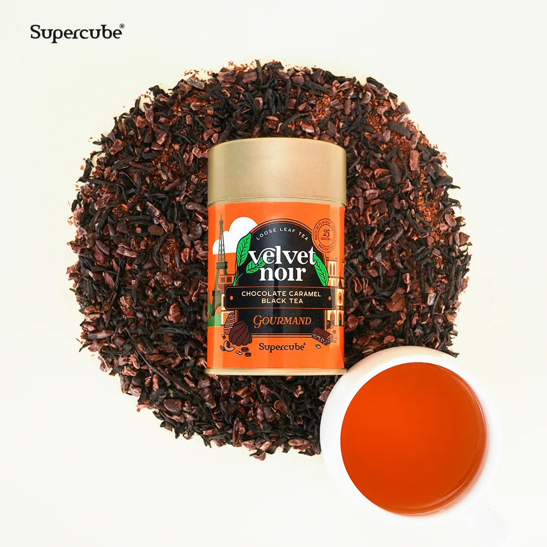 Supercube Velvet Noir Chocolate Caramel Black Tea tin on a bed of loose leaf tea and cocoa with a cup of brewed tea