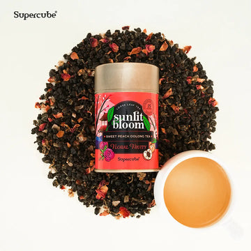 Supercube Sunlit Bloom Sweet Peach Oolong tea tin on a bed of loose leaf tea with a cup of brewed tea